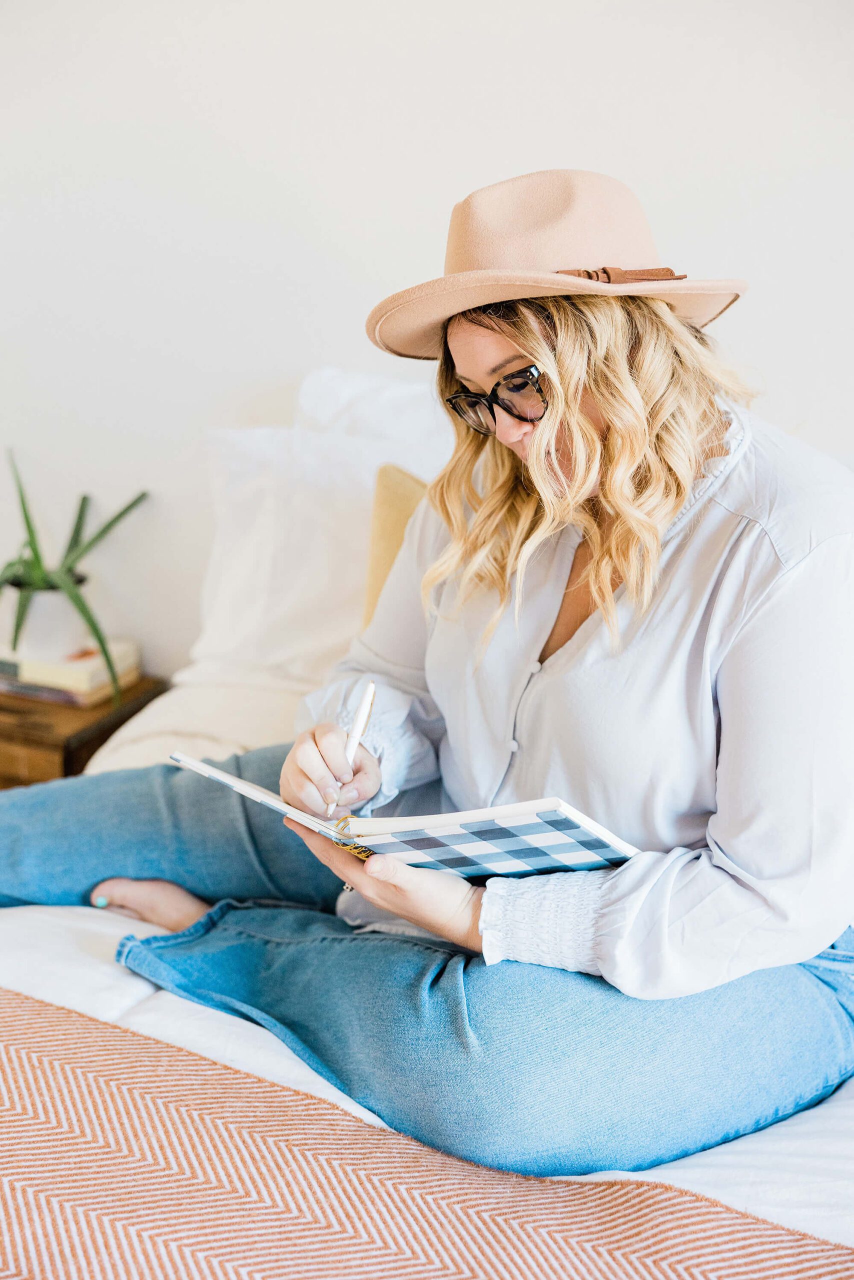 Blond woman in jeans and wide-brimmed hat writing in a blue notebook
